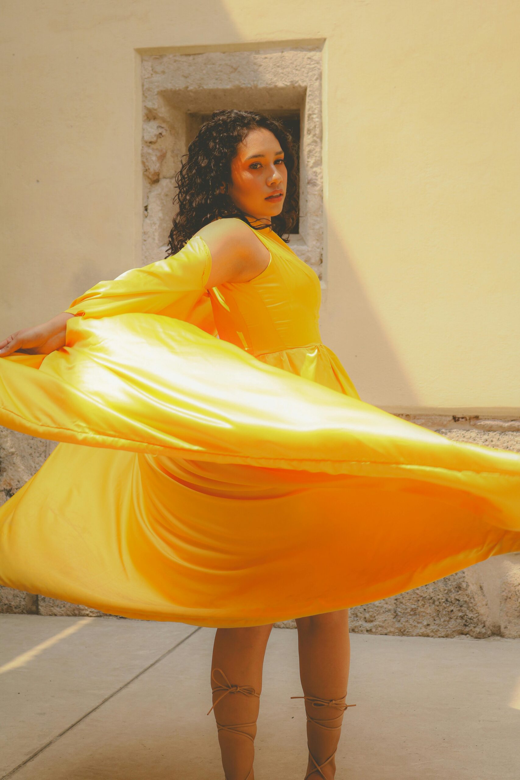A woman in a bright yellow flowing dress twirls gracefully in front of a sunlit wall, creating movement and light in the fabric.